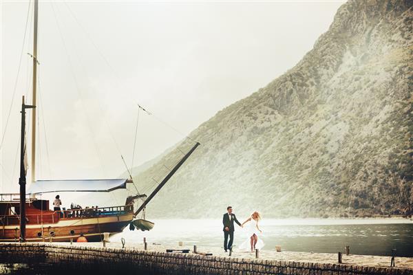 عروس و داماد جوان شیک در حال دویدن در اسکله نزدیک قایق تفریحی در پس زمینه دریا و کوههای مونته نگرو
