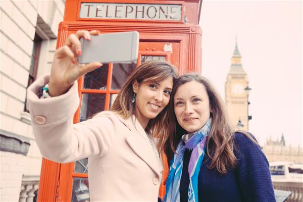 دو زن زیبا که در پس زمینه بیگ بن و غرفه تلفن قرمز در لندن سلفی می گیرند آنها بیست ساله هستند و تلفن را در دست دارند و به آن نگاه می کنند روی صورت تمرکز کنید
