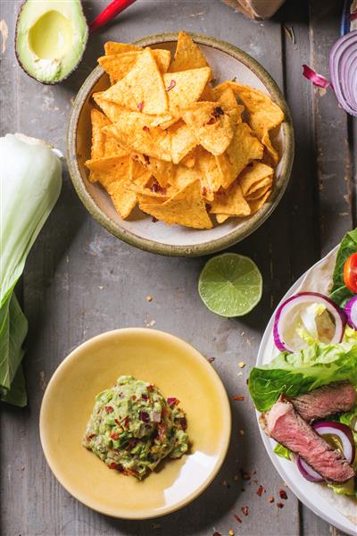 نمای بالایی از شام به سبک مکزیکی با تاکو سبزیجات چیپس ناچوس و گواکومول که روی میز چوبی خاکستری سرو می شود