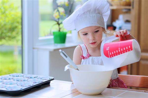 بچه کوچک خوشحال نوپای با کلاه آشپز سفید که آرد را به ظرف با مواد خمیر اضافه می کند و به مادر کمک می کند تا شیرینی خوشمزه ای در آشپزخانه تهیه کند