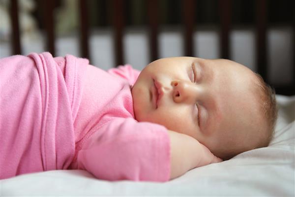 یک نوزاد دختر تازه متولد شده یک ماهه روی تختخواب خود روی پشت خوابیده و در یک پتوی صورتی حلقه شده است