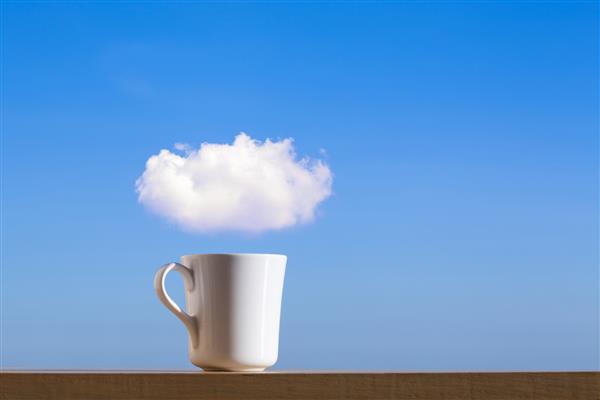 یک فنجان قهوه سفید روی میز چوبی روی آسمان آبی و ابرها
