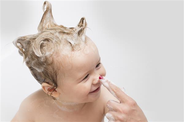 پسر بچه ای زیبا و مرطوب در اتاق حمام با موهای صابونی کف که نشسته و با مادر روی دست سفید و تصویر افقی بازی می کند