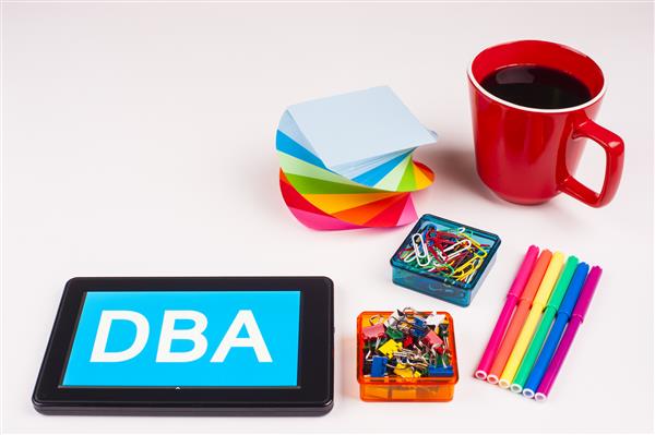 عبارت تجاری در رایانه لوحی - رنگهای رنگین کمانی رنگی فنجان دفترچه یادداشت خودکارها گیره های کاغذی سطح سفید - کلمات رنگ سفید در پس زمینه آبی - DBA