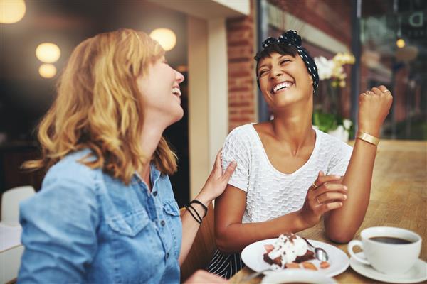 دو دوست دختر جوان و چند ملیتی که در کنار یکدیگر در یک رستوران از قهوه لذت می برند و در حالی که برای نشان دادن محبت لمس می کنند می خندند و شوخی می کنند