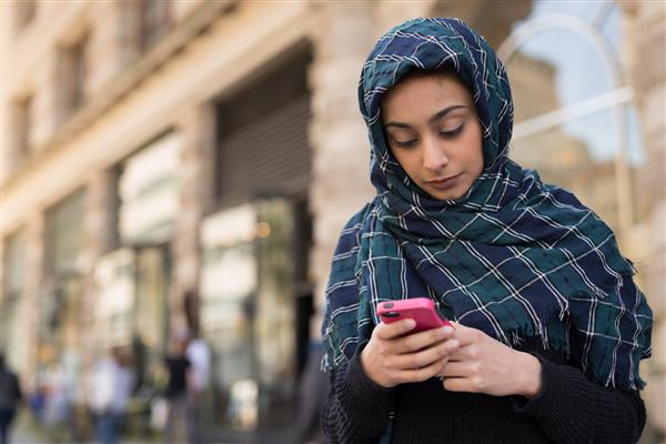 زن جوانی که روسری خود را با روسری حجاب پوشانده و در حال ارسال پیام کوتاه به تلفن همراه در شهر است