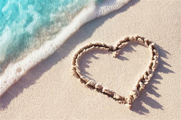 نماد قلب در شن و ماسه ساحل با موج نرم و آبی در پس زمینه