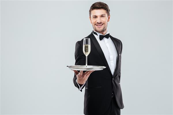 پیشخدمت در تاکسیدو با پاپیون ایستاده و به شما یک لیوان شامپاین را روی زمینه سفید ارائه می دهد