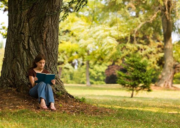 دختر دبیرستانی در حال خواندن کتاب در زیر درخت بزرگی در محیطی شبیه پارک