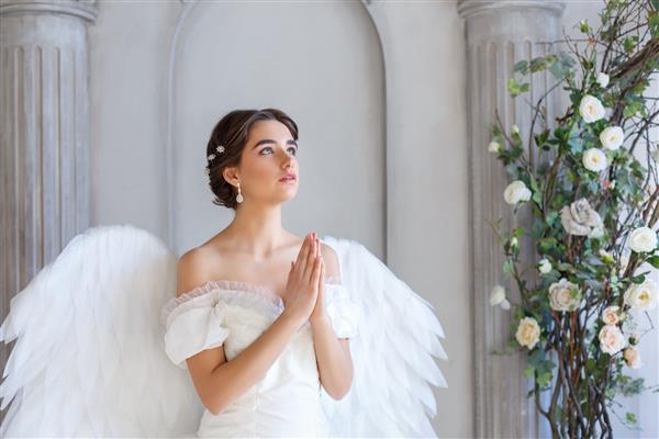 پرتره زن جوان زیبا با لباس سفید و بالهای فرشته ایستاده با ظاهری التماس آمیز
