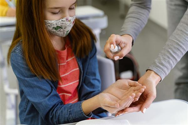 دختر بچه ای با ماسک پزشکی از معلم ضدعفونی کننده دست می گیرد