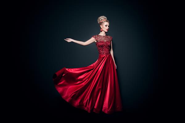 تصویر تمام قد از یک زن جوان فوق العاده که در لباس قرمز بلند و بلند با دست بلند بر روی زمینه مشکی پوشیده شده است