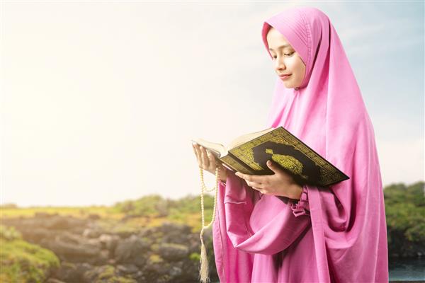 زن مسلمان آسیایی با حجاب مهره های نماز را در دست دارد و در فضای باز قرآن می خواند