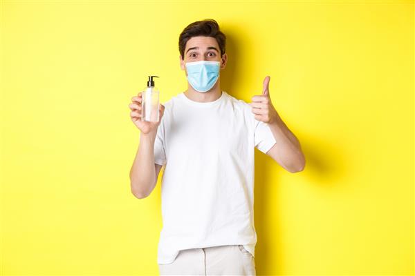مفهوم کووید -19 قرنطینه و شیوه زندگی مرد جوان راضی با ماسک پزشکی ضدعفونی کننده دست خوب نشان می دهد انگشت شست را بالا می آورد و زمینه ضد عفونی کننده زرد را توصیه می کند