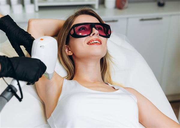 زن با لباس فرم با استفاده از دستگاه لیزر موهای زائد در اتاق با مشتری در کلینیک زیبایی روی میز پزشکی دراز کشیده است