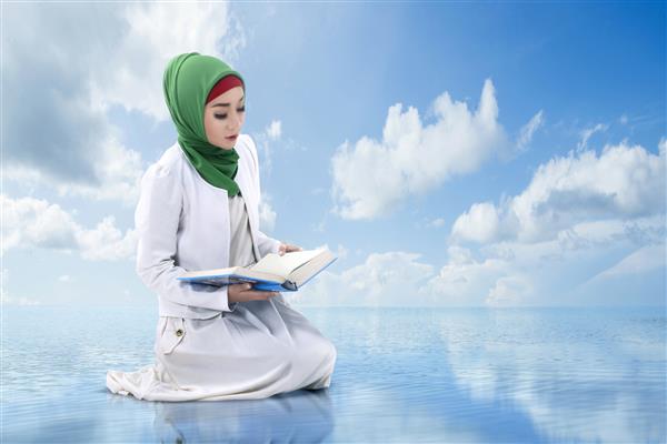 زن مسلمان آسیایی با حجاب نشسته و با آسمانی آبی قرآن می خواند