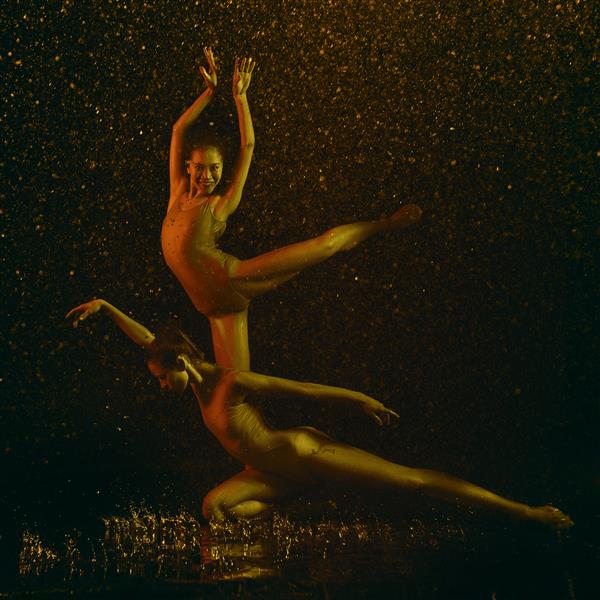 لبخند دو رقاص زن جوان باله زیر قطره آب و اسپری مدل های قفقازی و آسیایی با هم در چراغ های نئون می رقصند باله و مفهوم رقص معاصر عکس هنری خلاقانه