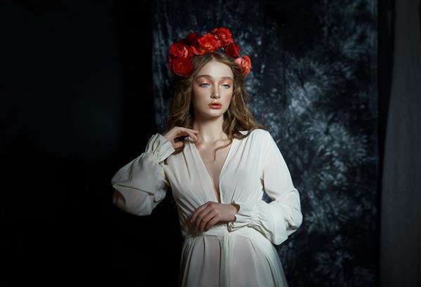 پرتره عاشقانه بهاری زن جوان بلوند با تاج گل از گل رز قرمز دختری با لباس سفید روشن یک زن با پوست کامل در پس زمینه تیره ژست می گیرد