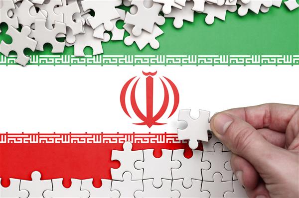پرچم ایران روی میزی به تصویر کشیده شده است که دست انسان روی آن یک پازل سفید رنگ جمع می کند
