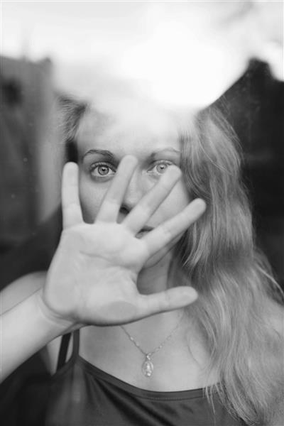عکس سیاه و سفید از زن جوان خوش تیپ با موهای روشن پشت شیشه و صورت خود را با دست می پوشاند و می گوید بس کنید