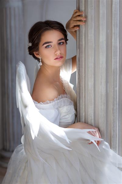 پرتره زن جوان زیبا با لباس سفید و بال فرشته ایستاده با ظاهری التماس آمیز در پس زمینه دیوار با ستون و گل