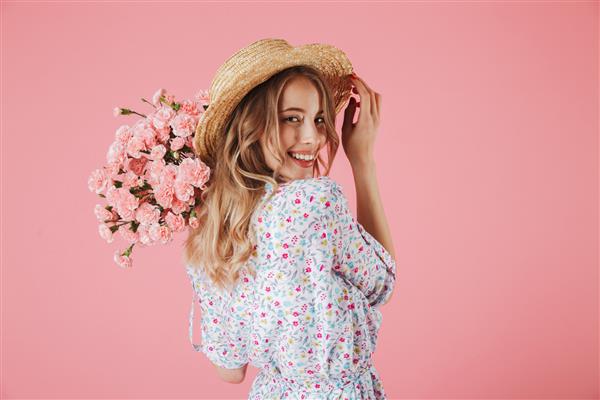 پرتره نزدیک از یک زن جوان جذاب با لباس تابستانی و کلاه حصیری که دسته گل میخک در دست دارد و روی شانه خود جدا شده روی پس زمینه صورتی نگاه می کند