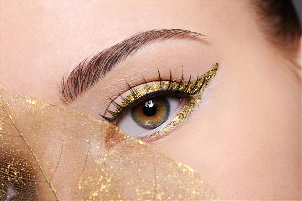 ماکرو شات از چشم زن زیبایی با خط چشم طلایی آرایشی روی برگ زرد مصنوعی را پوشانده است