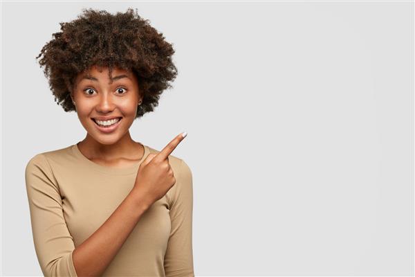 عکس افقی از زنی با پوست تیره زیبا با مدل موی آفرو لبخند پهن دندان‌های سفید چیزی زیبا را به دوست نشان می‌دهد به گوشه سمت راست بالا اشاره می‌کند مقابل دیوار می‌ایستد