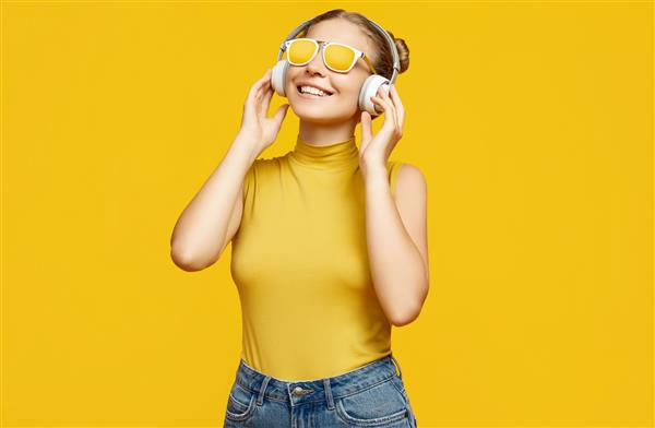 پرتره دختر خوشگل بلوند هیپستر با موهای مجعد که عینک آفتابی به چشم دارد از موسیقی در هدفون در پس زمینه زرد لذت می برد