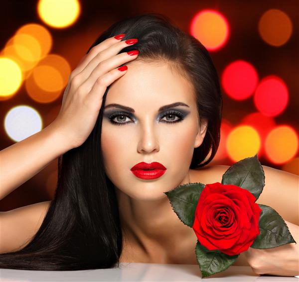 پرتره یک زن جوان زیبا با لب های قرمز ناخن ها و گل رز در دست مدل مد با آرایش چشم مشکی که در استودیو روی توپ های چراغ شب ژست گرفته است مفهوم پس زمینه نرم بوکه