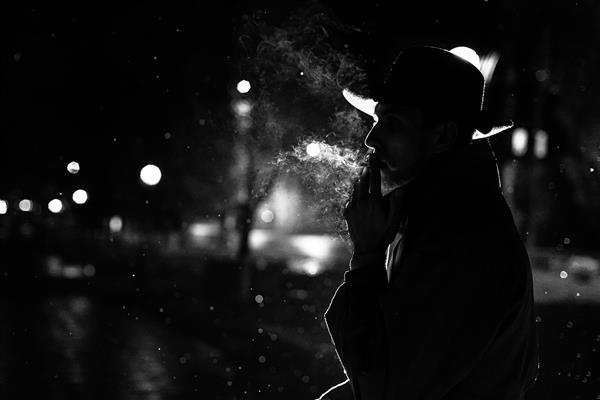 شبح تاریک مردی با کلاه در حال کشیدن سیگار زیر باران در خیابان شبانه به سبک نوآر