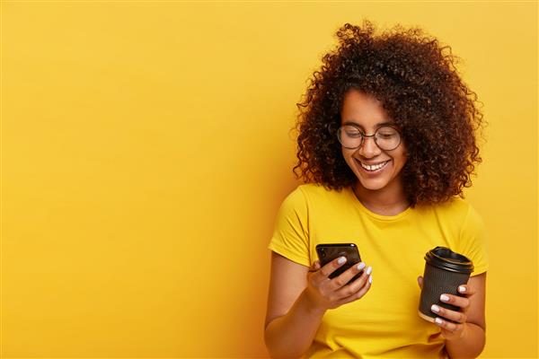 دختر نوجوان شاد با موهای مجعد موبایل مدرن در دست دارد قهوه بیرون بری دارد از طریق اپلیکیشن آنلاین تاکسی سفارش می دهد پیامک می نویسد لباس زرد می پوشد مردم سبک زندگی مدرن و تکنولوژی