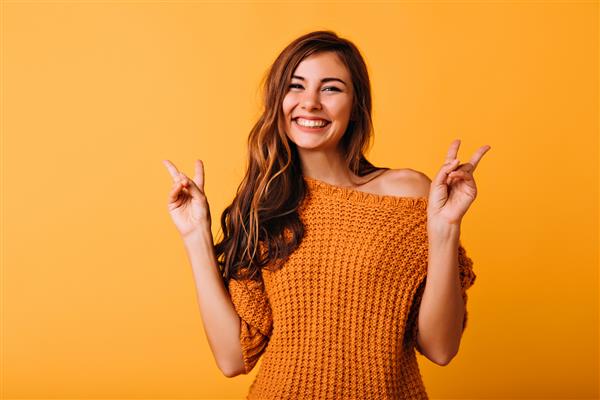 دختری دلربا با مدل موی بلند که با علامت صلح روی نارنجی ژست گرفته است مدل زن خوشحال با ژاکت بافتنی مد روز در حال خندیدن در استودیو