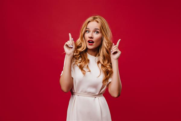 زن جوان علاقه مند با موهای موج دار بلند که با دهان باز ژست گرفته است دختر شیک دبونیر با لباس سفید روی دیوار قرمز ایستاده است