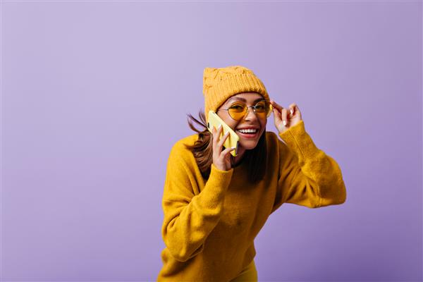 دختر 21 ساله شاد با موهای قهوه ای رنگ بامزه و جالب با تلفن صحبت می کند زنی خندان که از عینک آفتابی زرد با ژست گرفتن برای پرتره نگاه می کند