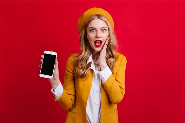 زن پر زرق و برق متعجب که گوشی هوشمند با صفحه نمایش خالی روی دیوار قرمز در دست دارد دختر بلوند جذاب با کلاه زرد با دستگاه دیجیتالی
