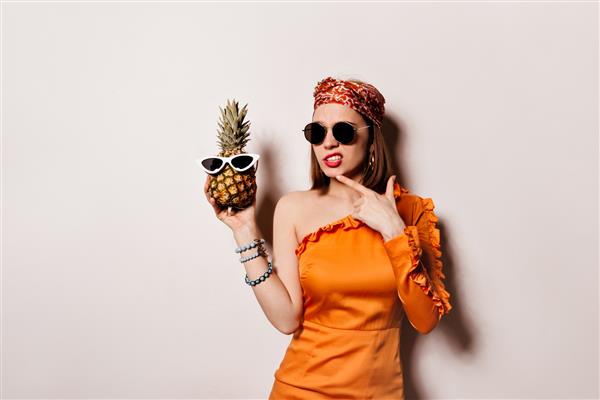 خانمی با لباس نارنجی و عینک آفتابی متفکرانه ژست گرفته و آناناس را در فضایی منزوی گرفته است