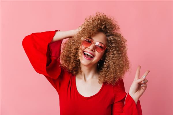 زن بلوند شاد با فر می خندد و علامت صلح را در فضای صورتی نشان می دهد عکس فوری از خانمی با عینک آفتابی و تاپ قرمز