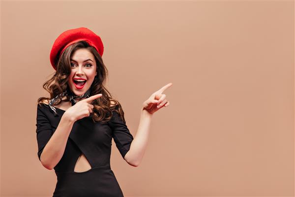 دختر زیبا با موهای موج دار لبخند می زند و با انگشتانش به سمت راست اشاره می کند پرتره زنی با کلاه قرمز و لباس مشکی شیک در زمینه بژ