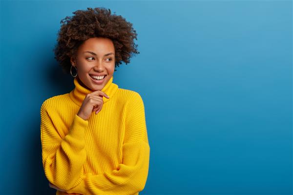 زن جوان مجعد مثبت با ژاکت راحت زرد رنگ چانه را در دست گرفته با حالتی رویایی به کناری نگاه می کند ایده جالبی در ذهن دارد روی پس زمینه آبی جدا شده است