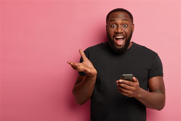 عکس افقی از مرد سیاهپوست شاد با استفاده از تلفن همراه مدرن ژست های دست فریاد زدن از احساسات مثبت دریافت پیام خوب پوشیدن تی شرت مشکی