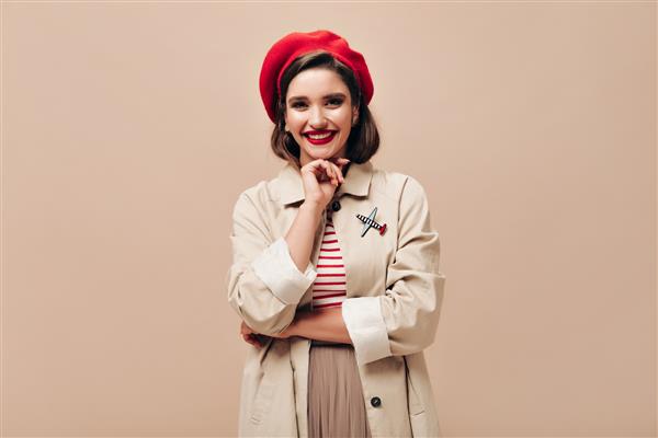 بانوی زیبا با کلاه قرمز و لبخند در زمینه بژ زن جوان شادی با کلاه قرمزی و کت روشن به دوربین لبخند می زند