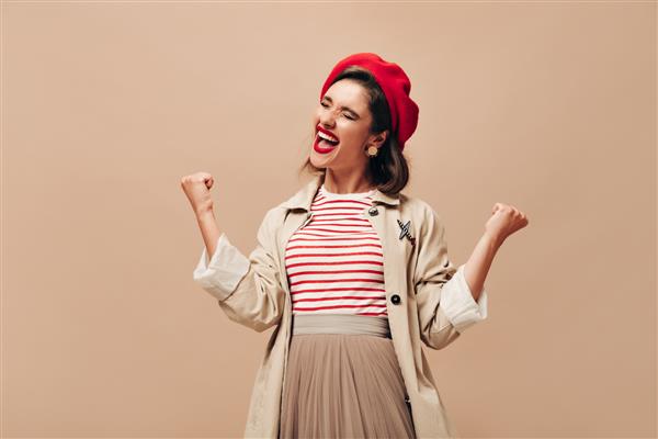 خانمی با سنگر و کلاه مد روز که به صورت احساسی روی پس زمینه بژ ژست گرفته است زن جوان شاد با موهای تیره در کلاه قرمزی شادی می کند