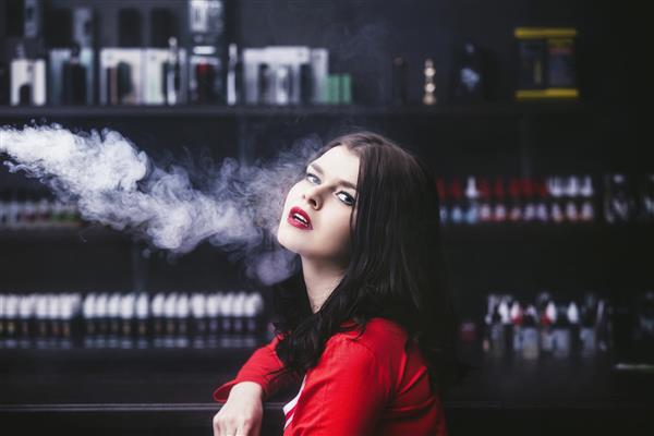 زن جوان سبزه زیبا با آرایش مد در بار با بخاری از سیگار الکترونیکی