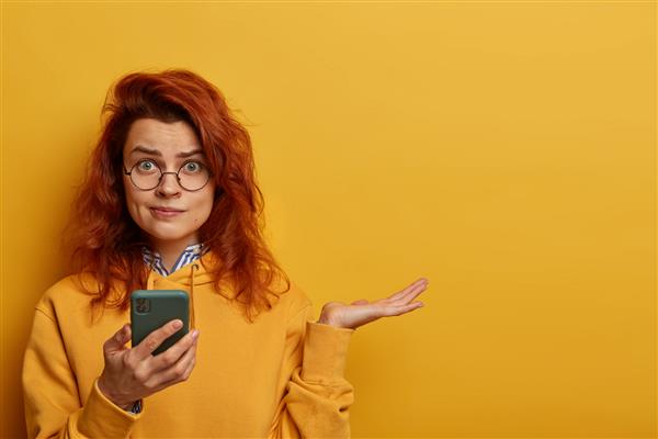 زن مو قرمز متحیر خشمگین کف دستش را بالا می‌گیرد فکر می‌کند در پیام دریافتی چه پاسخی بدهد تلفن همراه در دست دارد عینک گرد و هودی می‌پوشد مدل‌هایی روی دیوار زرد با فضای خالی درست است