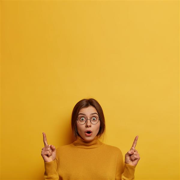 زن جوان شوکه شده در مورد آخرین اخبار شایعات می کند هر دو انگشت اشاره را به سمت بالا نشان می دهد اخبار غافلگیرکننده ای می شنود دهان باز می کند عینک گرد و یقه اسکی بزرگ می پوشد روی دیوار زرد جدا شده است