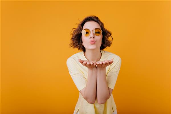زن عاشقانه مو کوتاه با عینک قدیمی که با حالت چهره دوست داشتنی ژست گرفته است دختر شادی با تی شرت زرد در حال ارسال بوسه هوایی در هنگام عکاسی
