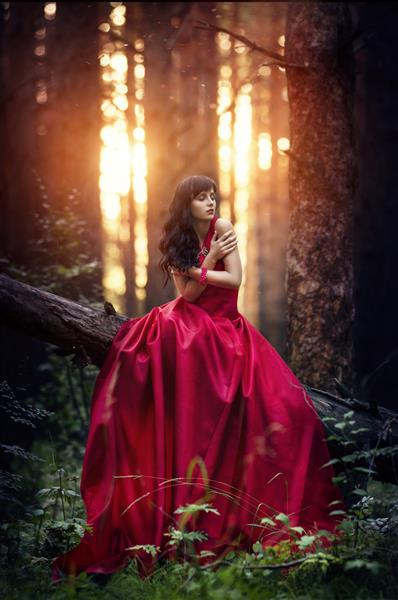 زنی با لباس قرمز بلند به تنهایی در جنگل تصویر افسانه ای و مرموز از یک دختر در یک جنگل تاریک در آفتاب عصر غروب خورشید در جنگل