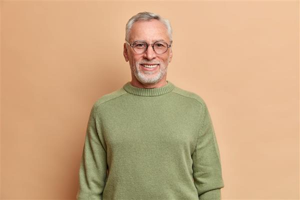 عکس نیم قد از مرد سالخورده شادی که با دندان های سفید لبخند می زند با عینک نوری و ژاکت جدا شده روی دیوار قهوه ای