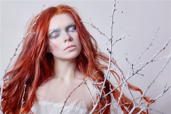 دختر مو قرمز با موهای بلند صورت پوشیده از برف با یخ ابروها و مژه های سفید در یخبندان شاخه درخت پوشیده از برف ملکه برف و زمستان آرایش زمستانی صورت زن سر قرمز